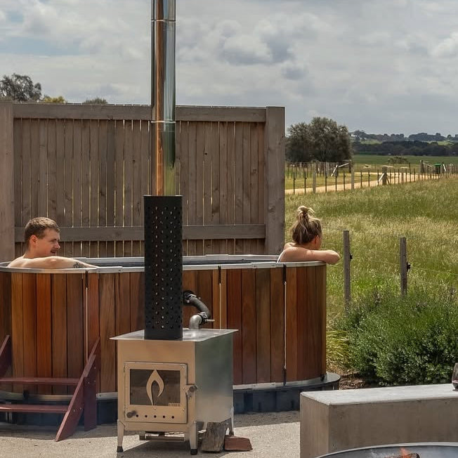 Two people in a hot tub with a view of a field and a fire pit.